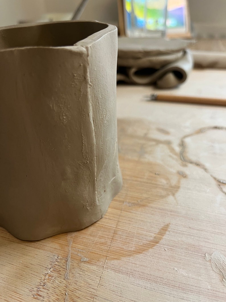 May include: A close-up of a light brown ceramic pot with a textured surface. The pot is sitting on a wooden table.