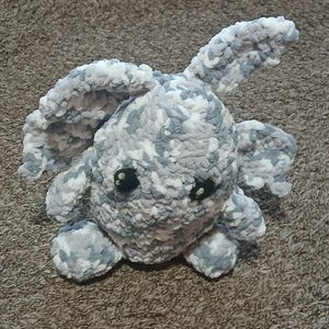 May include: A handmade, plush, crocheted bunny in shades of gray and white. The bunny has large, floppy ears, small arms and legs, and black safety eyes. The soft, textured yarn gives the bunny a cuddly appearance.