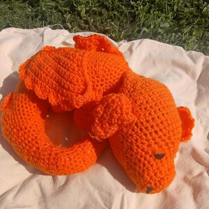May include: An orange crocheted dog toy, lying on a white cloth. The dog has floppy ears and a curled tail. The toy is made of a thick yarn and has a dark brown nose and eyes. The background is green grass.
