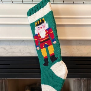 May include: A green knitted Christmas stocking with a white cuff and a nutcracker design. The nutcracker is wearing a red and yellow uniform with a black hat and a white beard.