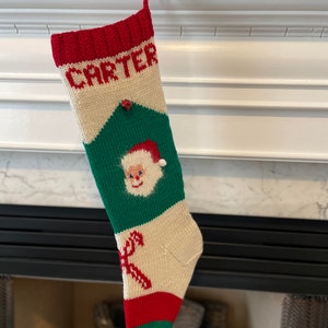 May include: A knitted Christmas stocking with a red, white and green colour scheme. The stocking has the name "CARTER" knitted into the top and a Santa Claus face in the middle.