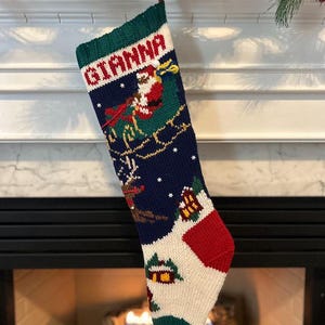 Puede incluir: Un calcetín de Navidad tejido con el nombre "GIANNA" en rojo y blanco. El calcetín presenta una escena de Papá Noel en su trineo, una casa y un diseño rojo y blanco. El calcetín está colgado sobre una chimenea.