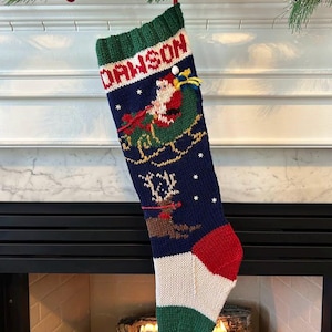 Puede incluir: Un calcetín navideño azul marino con el nombre "DAWSON" tejido en blanco. Se representa a Papá Noel en un trineo, junto con un reno. La punta es roja, el talón es blanco y el puño es verde.