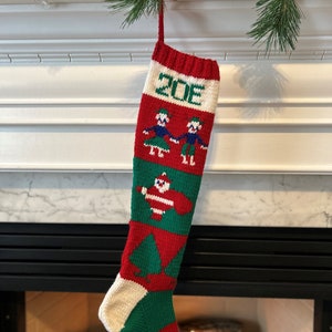 May include: A red and green knitted Christmas stocking with a white cuff and the name "ZOE" embroidered on the front. The stocking features a pattern of a couple holding hands, a snowman, and Christmas trees.