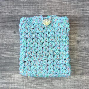 May include: A rectangular crochet pouch made from light blue yarn with subtle flecks of pink, yellow, and green throughout. The pouch features a thick, textured knit pattern and is secured at the top center with a single round, patterned button.