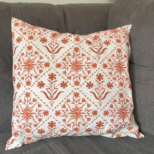 May include: Square throw pillow with a white background and a repeating pattern of orange floral designs. The pillow is resting on a gray sofa, showcasing its decorative appeal and potential for home decor.