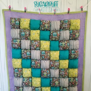 May include: A purple and green quilted baby blanket with a padded texture. The blanket is decorated with a repeating pattern of owls in various colors and designs.