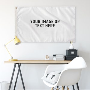 May include: A white rectangular flag with the text "YOUR IMAGE OR TEXT HERE" in black. The flag is hanging on a white wall above a wooden desk. A yellow desk lamp, a white chair, and other office supplies are also visible.
