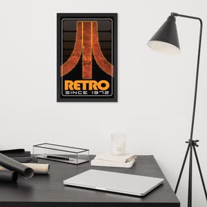 May include: A framed rectangular wall art print featuring a black background with a stylized orange and brown graphic logo resembling a mountain peak. Below the graphic, the text reads RETRO in large orange letters and SINCE 1972 in white.
