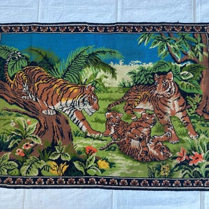 May include: A rectangular woven tapestry featuring a jungle scene with two adult tigers and two tiger cubs. One tiger rests on a tree branch while the others play in the grass. The border is dark blue with a repeating floral pattern and fringe.