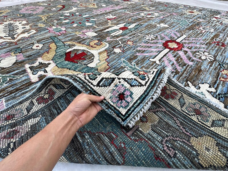 May include: A large area rug featuring a dark brown base with intricate floral and geometric patterns in shades of blue, red, pink, and cream. The rug has a visible pile texture and a fringed edge, with a corner folded over to reveal the underside.