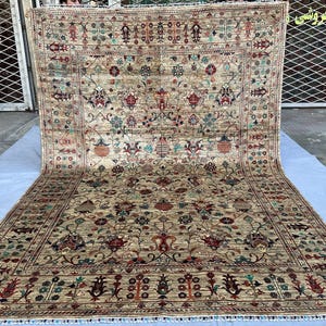 May include: A large rectangular area rug featuring a beige background with an intricate floral and vine pattern in shades of red, blue, green, and cream. The rug has a detailed border and a fringe along the bottom edge, displaying symmetrical motifs.