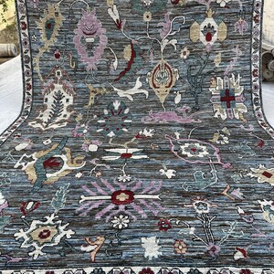 May include: A rectangular area rug featuring a blue and brown striated background with a variety of stylized floral and geometric motifs in shades of pink, cream, green, and red. The rug is framed by a decorative border with repeating floral patterns.