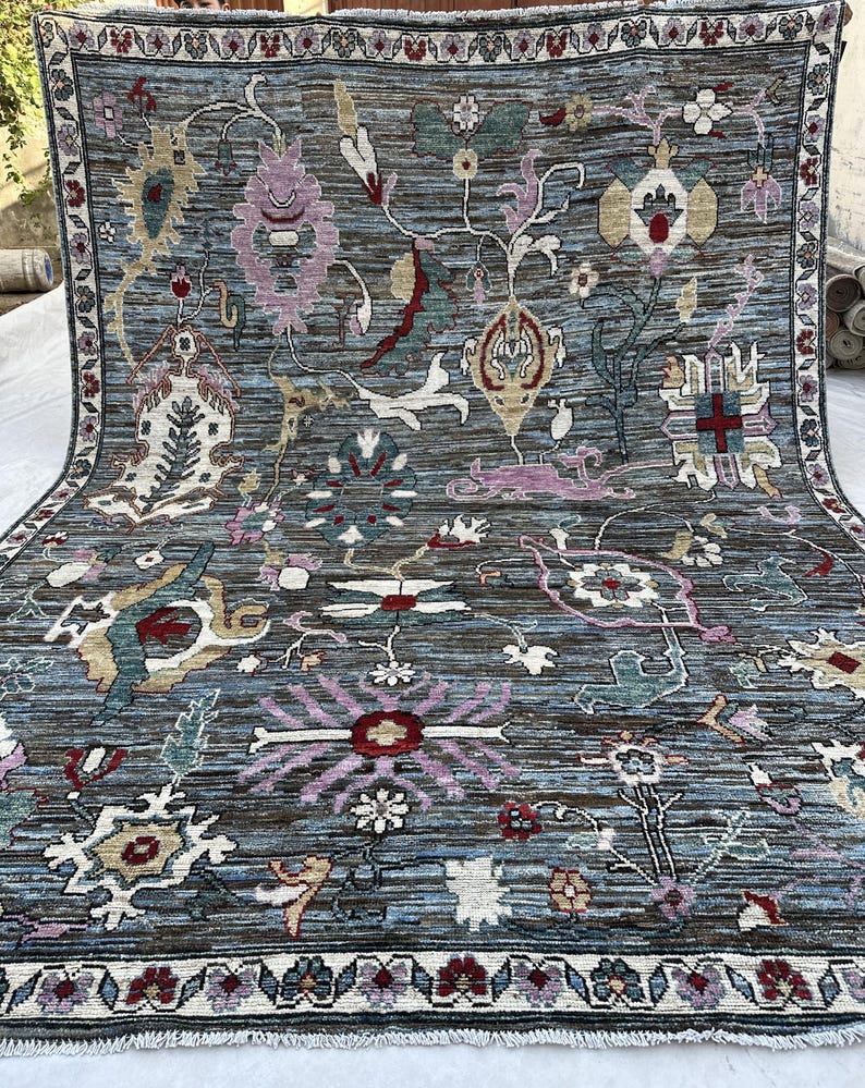 May include: A rectangular area rug featuring a blue and brown striated background with a variety of stylized floral and geometric motifs in shades of pink, cream, green, and red. The rug is framed by a decorative border with repeating floral patterns.