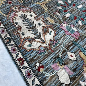 May include: A woven area rug featuring a blue and brown textured background with intricate floral and botanical patterns in cream, pink, teal, and red. The rug has a detailed border with small flower motifs and a dark, defined edge.