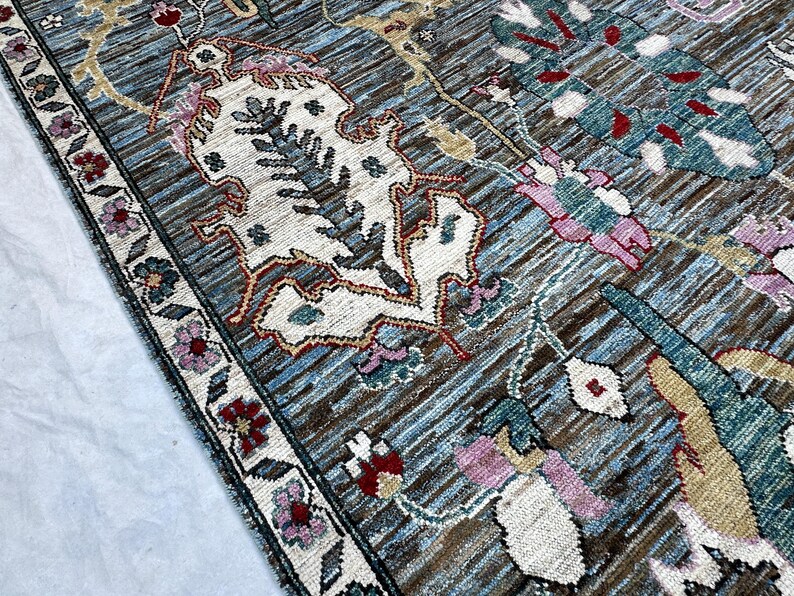 May include: A woven area rug featuring a blue and brown textured background with intricate floral and botanical patterns in cream, pink, teal, and red. The rug has a detailed border with small flower motifs and a dark, defined edge.