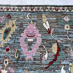 May include: A rectangular area rug featuring a blue and brown textured background with a repeating floral pattern. The design includes large pink, green, and cream-colored stylized blossoms and vines, framed by a border with small floral motifs.