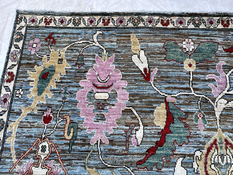 May include: A rectangular area rug featuring a blue and brown textured background with a repeating floral pattern. The design includes large pink, green, and cream-colored stylized blossoms and vines, framed by a border with small floral motifs.