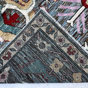 May include: A woven rug featuring a complex pattern with shades of blue, beige, red, and pink. The design includes floral motifs and geometric borders. One corner of the rug is folded over, revealing the textured underside and the intricate weave.