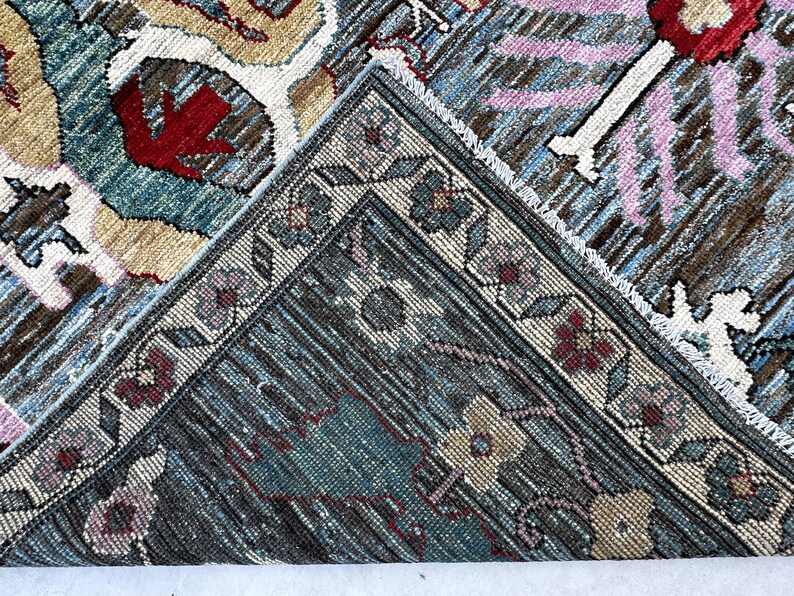 May include: A woven rug featuring a complex pattern with shades of blue, beige, red, and pink. The design includes floral motifs and geometric borders. One corner of the rug is folded over, revealing the textured underside and the intricate weave.