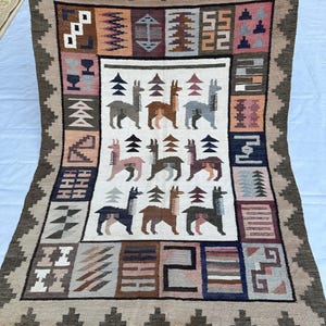May include: A rectangular woven rug featuring a central white panel with nine stylized llama figures arranged in three rows, surrounded by a wide border of geometric patterns in shades of brown, pink, blue, and cream, finished with fringe.