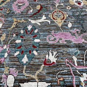 May include: A hand-knotted area rug featuring a complex floral and botanical pattern in shades of blue, brown, pink, white, and red. The design includes stylized blossoms, scrolling vines, and abstract shapes woven into a textured surface.