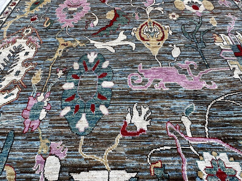 May include: A hand-knotted area rug featuring a complex floral and botanical pattern in shades of blue, brown, pink, white, and red. The design includes stylized blossoms, scrolling vines, and abstract shapes woven into a textured surface.