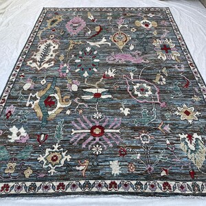 May include: A rectangular area rug featuring a dark blue and brown striated background with a complex pattern of stylized floral and botanical motifs in shades of pink, teal, cream, and red, finished with a decorative border and fringe.