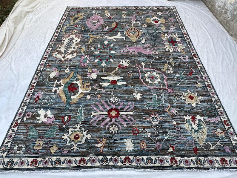 May include: A rectangular area rug featuring a dark blue and brown striated background with a complex pattern of stylized floral and botanical motifs in shades of pink, teal, cream, and red, finished with a decorative border and fringe.