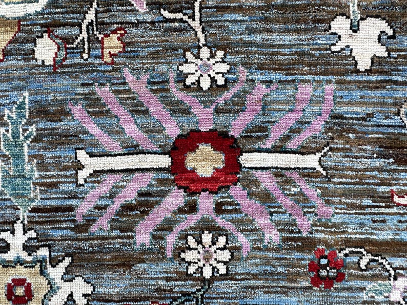 May include: A woven rug featuring a dark brown and blue textured background with a central floral motif in red, white, and light purple. The design includes repeating white flower shapes and intricate botanical patterns across the surface.