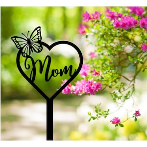 Puede incluir: Estaca de jardín de metal negro en forma de corazón con la palabra "Mamá" en el interior y una silueta de mariposa en la parte superior. La estaca está delante de un fondo borroso de follaje verde y flores rosas.