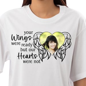 Puede incluir: Camiseta blanca con un gráfico negro de un corazón con alas de ángel. El corazón tiene una foto de una persona dentro. El texto dice "tus alas estaban listas pero nuestros corazones no lo estaban".