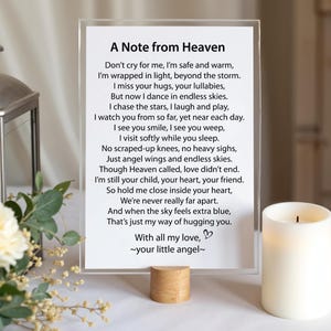 May include: A rectangular clear acrylic sign featuring the printed poem A Note from Heaven in black text, supported by a cylindrical light wood base. The sign is positioned next to a white pillar candle and a bouquet of white flowers.