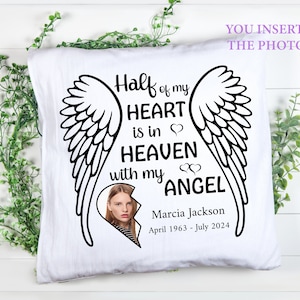 Op de afbeelding: Wit vierkant kussen met een zwarte afbeelding van engelenvleugels en de tekst "Half of my HEART is in HEAVEN with my ANGEL." Bevat een foto en de naam "Marcia Jackson" met data "April 1963 - juli 2024."