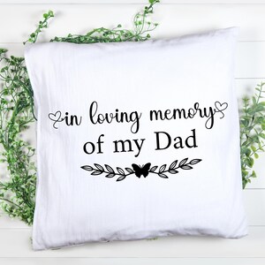 Peut inclure: Coussin carré blanc avec l'inscription noire "in loving memory of my Dad". Le texte est orné de cœurs, de feuilles et d'un papillon. Des brins de verdure entourent le coussin, sur fond de bois blanc.