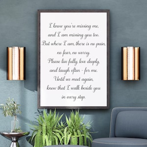 Op de afbeelding: Een rechthoekige wanddecoratie in een donkerhouten lijst met zwarte cursieve tekst op een witte achtergrond: I know you're missing me, and I am missing you too. But where I am, there is no pain, no fear, no worry. Please live fully, love deeply, and laugh often - for me. Until we meet again, know that I walk beside you in every step.