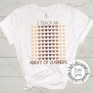 May include: White t-shirt with the text "I TEACH AN ARRAY OF LEARNERS" in brown font. The design features a grid of heart shapes in various shades of brown and tan. A circular logo is in the bottom right corner.