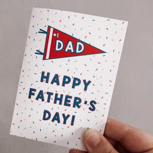 May include: A Father's Day greeting card held by a hand. The card features a red pennant with the word "DAD" and the text "HAPPY FATHER'S DAY!" in blue. The background is white with scattered confetti.