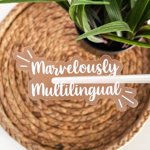 May include: A clear sticker with the text "Marvellously Multilingual" in white lettering on a brown background. The sticker is placed on a woven straw surface.