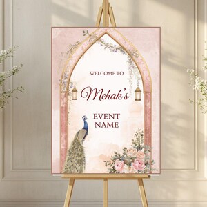 May include: A rectangular event welcome sign on a wooden easel featuring a gold archway, a peacock, and floral arrangements. The text reads WELCOME TO Mehak's EVENT NAME. The background is a soft, pale pink with hanging vines and two gold lanterns.