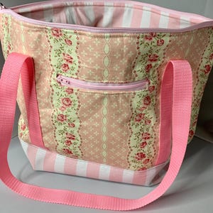 May include: A pink tote bag with a floral and striped pattern. The bag features a pink zipper pocket and pink straps. The interior lining is white and pink striped. The bag is approximately 14 inches wide and 12 inches tall.