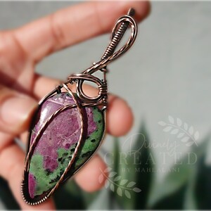 May include: A handcrafted pendant featuring a teardrop-shaped ruby zoisite gemstone wrapped in copper wire. The stone displays a mix of deep pink and green hues. The pendant is suspended from a copper wire loop.