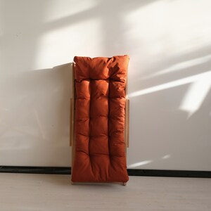 May include: A wooden folding chair with a burnt orange cushion. The chair is folded and standing upright.