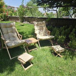 May include: Two wooden chairs with beige cushions and a small wooden table with a beige cushion footrest in a grassy garden setting.