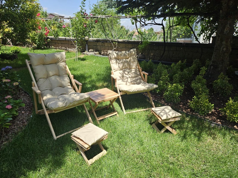 May include: Two wooden chairs with beige cushions and a small wooden table with a beige cushion footrest in a grassy garden setting.