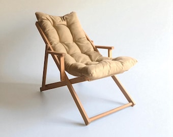 May include: A folding chair constructed from light-toned wood featuring a tufted, beige-colored cushion that covers the backrest and seat. The chair has a simple frame design with armrests and a reclining back position for support.