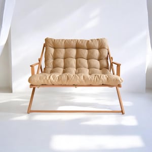 May include: A light brown wooden folding loveseat with a beige padded cushion. The loveseat is in a white room with a white curtain in the background.