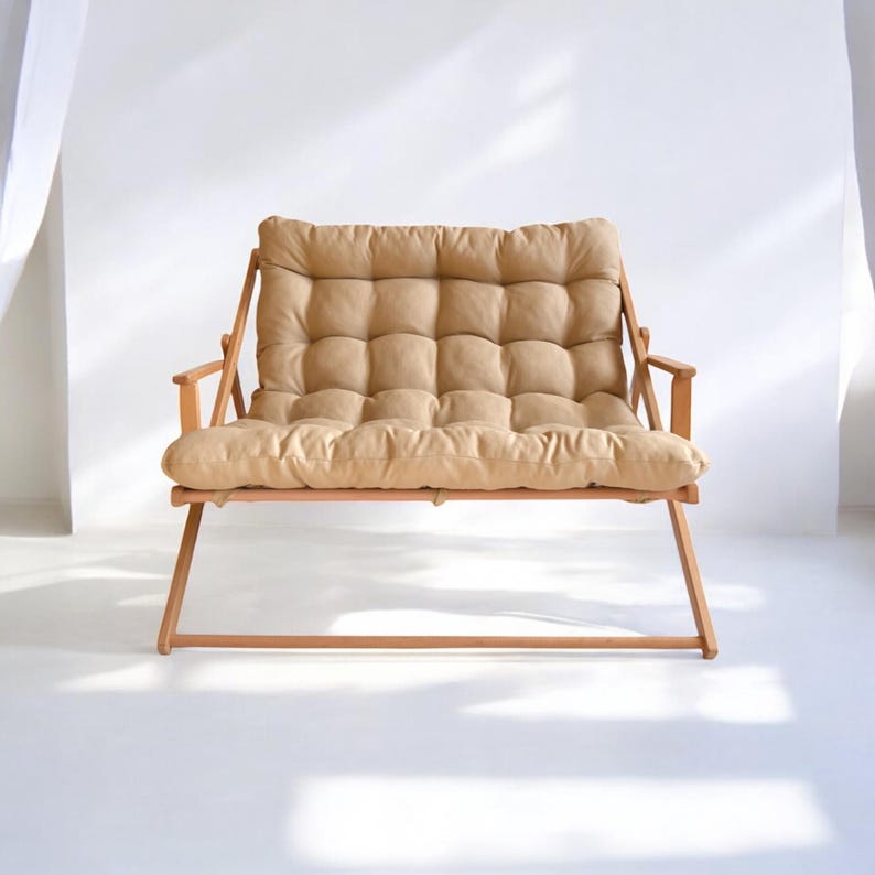 May include: A light brown wooden folding loveseat with a beige padded cushion. The loveseat is in a white room with a white curtain in the background.