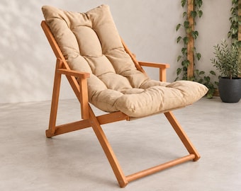 May include: A folding lounge chair constructed from light-toned wood, featuring a tufted, beige-colored cushioned seat and backrest. The chair has a simple frame design with armrests and a slanted back, providing a reclined seating position.