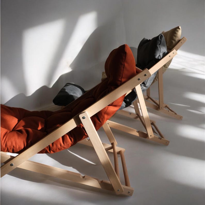 May include: Three wooden folding chairs with fabric cushions. The chair in the foreground has an orange cushion, the middle chair has a gray cushion, and the chair in the background has a beige cushion.